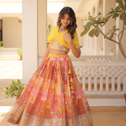 Woman in a yellow top and colorful skirt standing in an outdoor setting with plants and architectural elements.